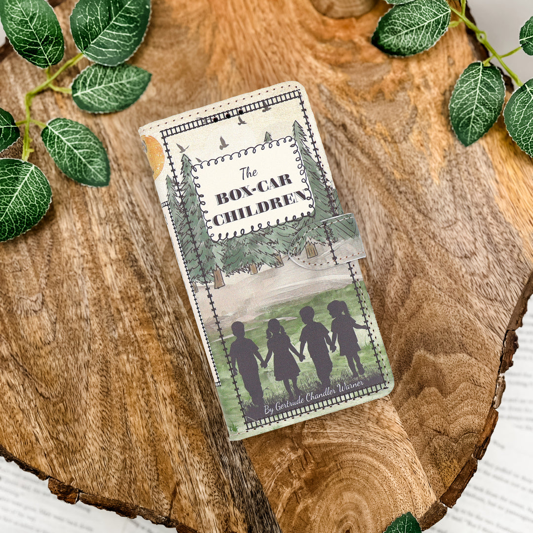 Book phone case inspired by 'The Boxcar Children' on a wooden surface with green leaves.