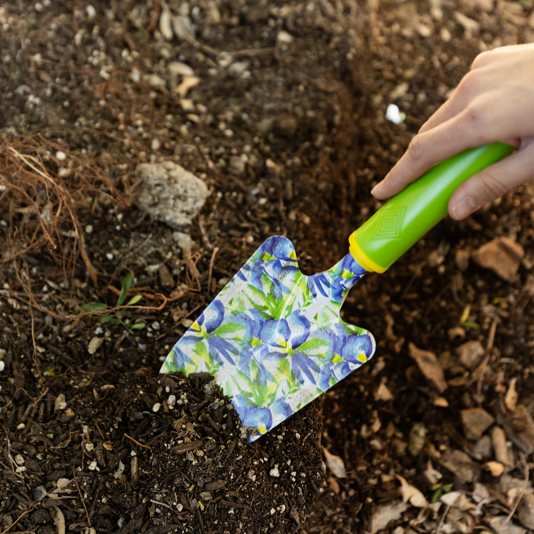 A hand holds a lime green gardening trowel with blue floral pattern, digging into the soil