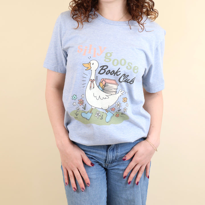 A woman wearing a light blue t-shirt with a Silly Goose Book Club graphic and text on a beige background