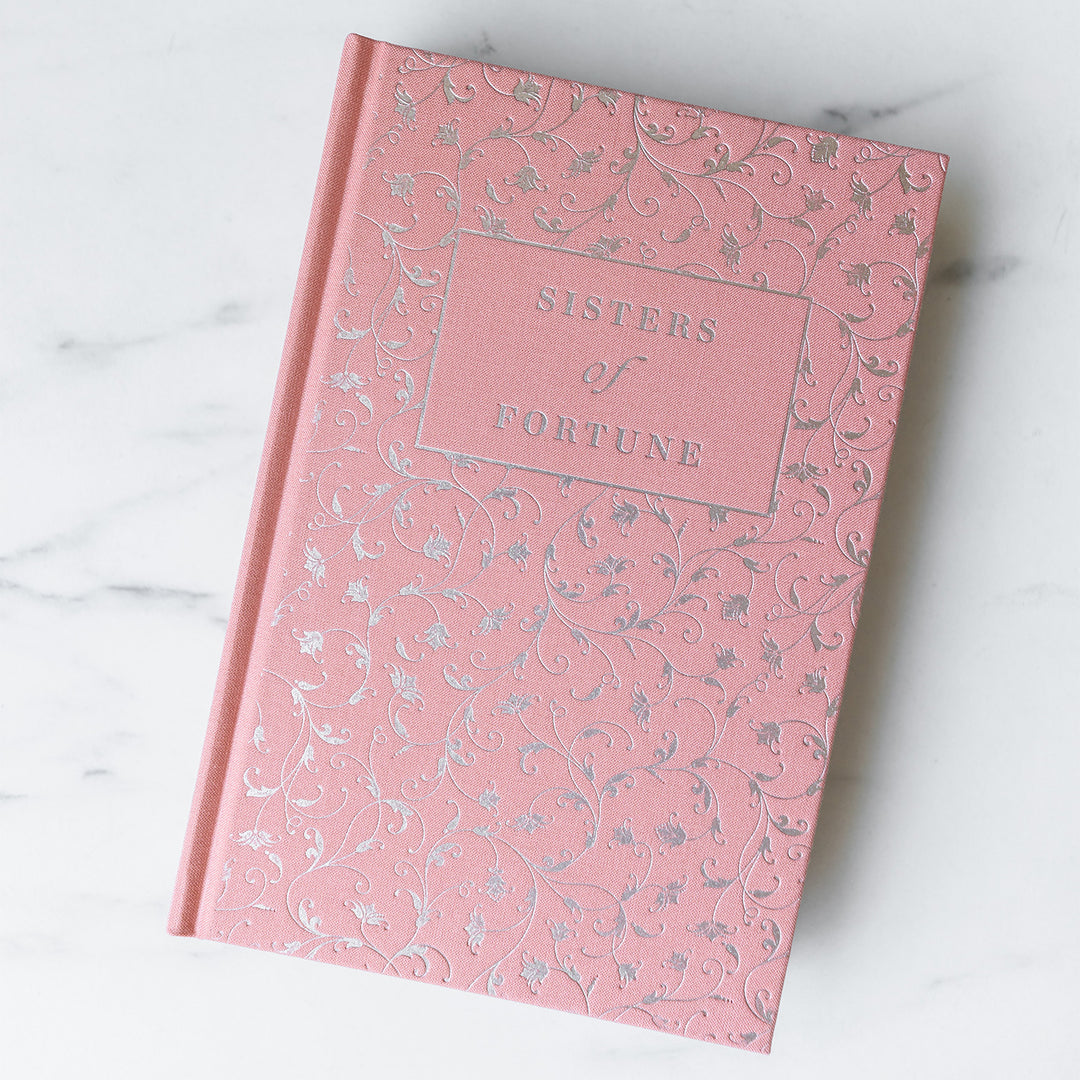 An exclusively designed hardcover copy of Sisters of Fortune by Anne Lee Huber as designed by the Once Upon a Book Club team. The hard case is light pink and features silver foiling with floral filigree.