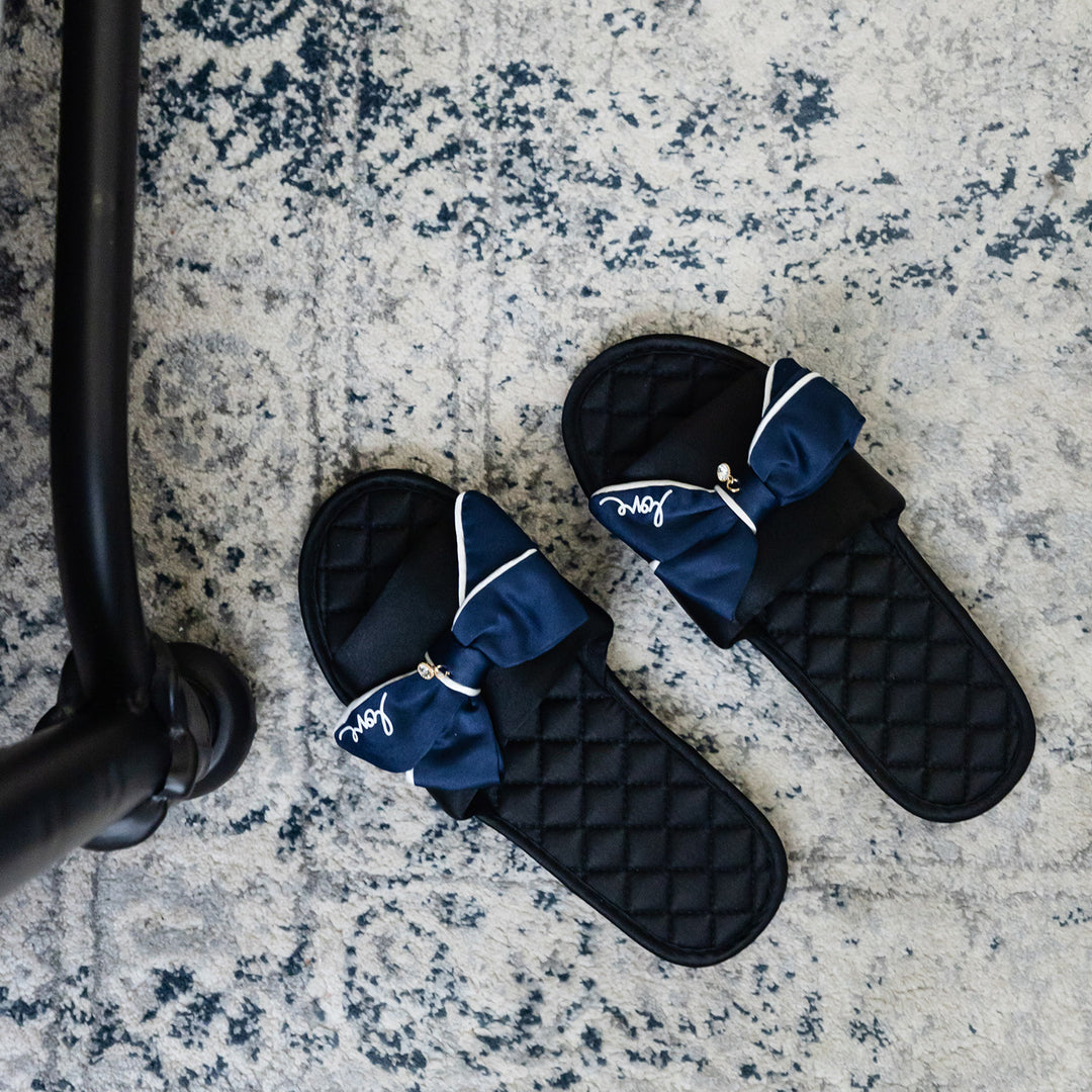 a pair of black and navy blue slippers with the word 'love' printed on the navy blue bow. a crystal hangs from the blue center bow. they sit on an ornate rug.