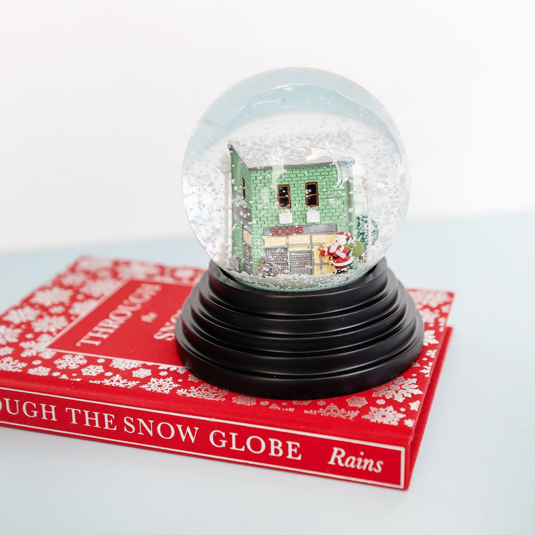 a close up of a custom designed snowglobe featuring a green brick bookstore with Santa outside. The snowglobe sits on top of the fabric bound hardcover edition of Through the Snow Globe by Annie Rains.