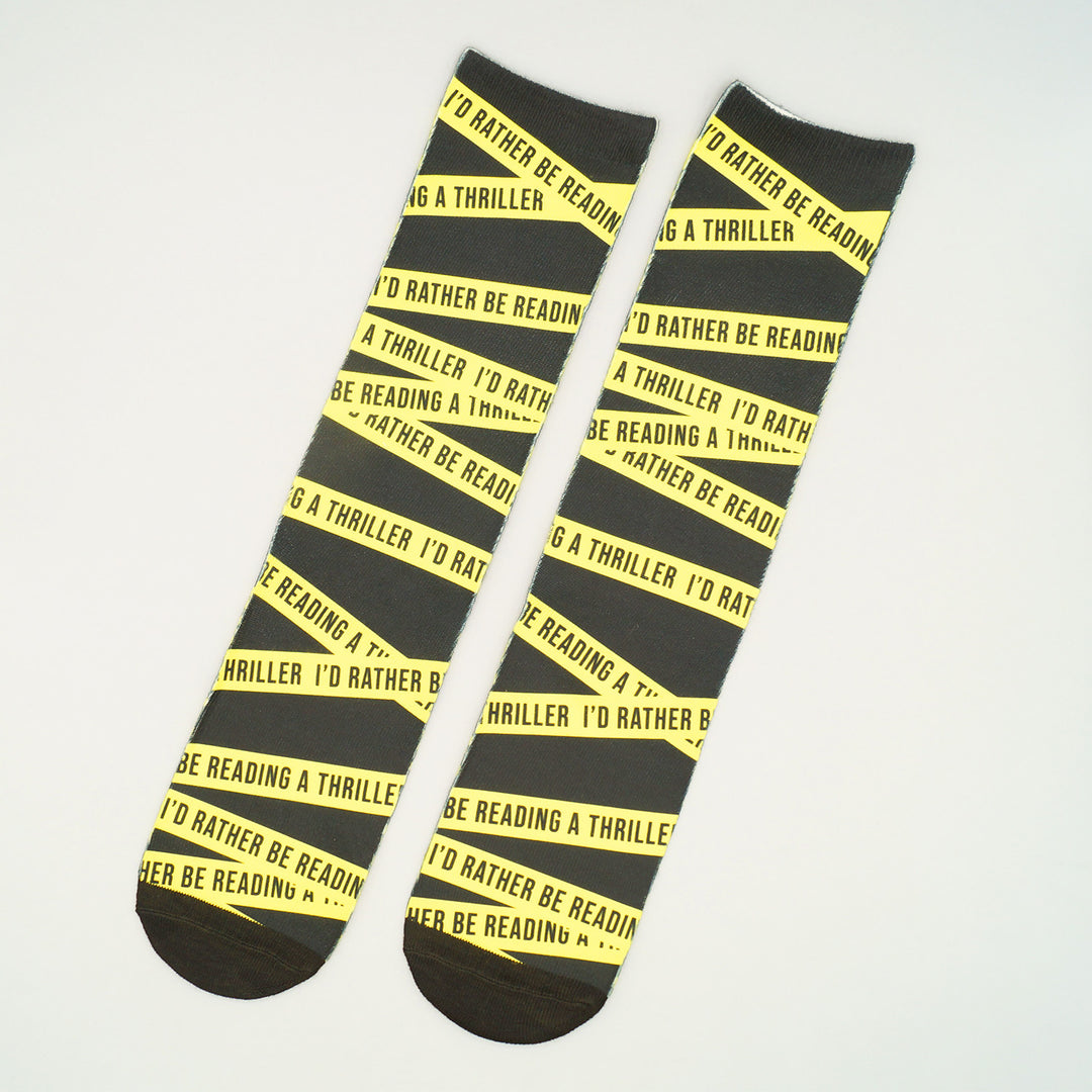 A pair of black long socks with yellow crime scene tape pattern. On the crime scene tape, it says I'd Rather Be Reading a Thriller repeated.