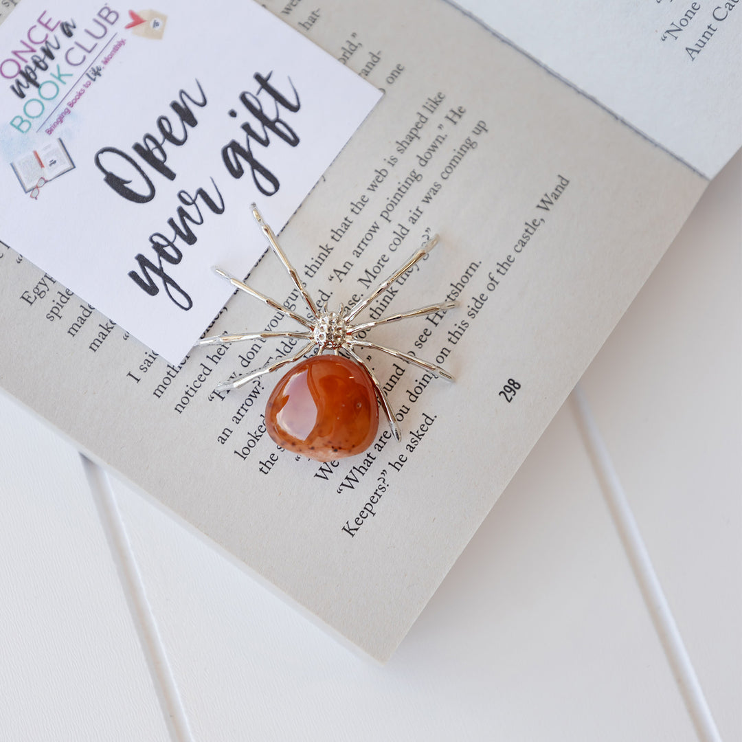 A silver spider figure on an open book with an orange quartz gemstone for it's body.
