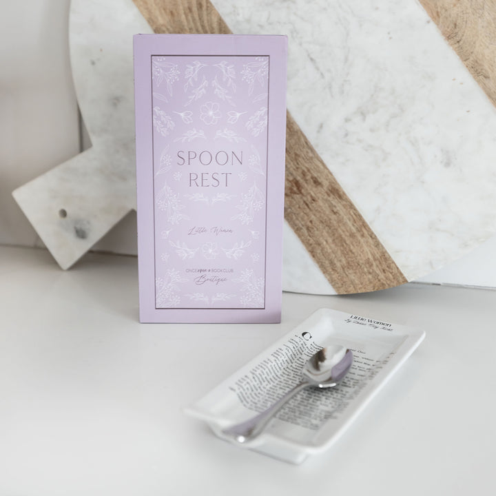 A purple magnetic book-shaped box labeled "Spoon Rest" "Little Women" "Once Upon a Book Club Boutique" stands toward the back of the countertop against a marble and wood serving tray. The spoon rest lays toward the front of the image holding a spoon.