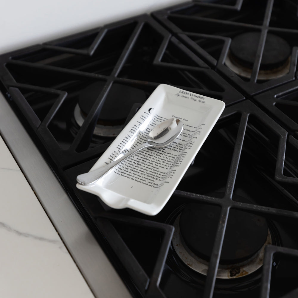 A ceramic spoon rest inspired by Little Women sits on a gas stovetop holding a spoon. The book page printed on the spoon rest is Chapter One of the novel.