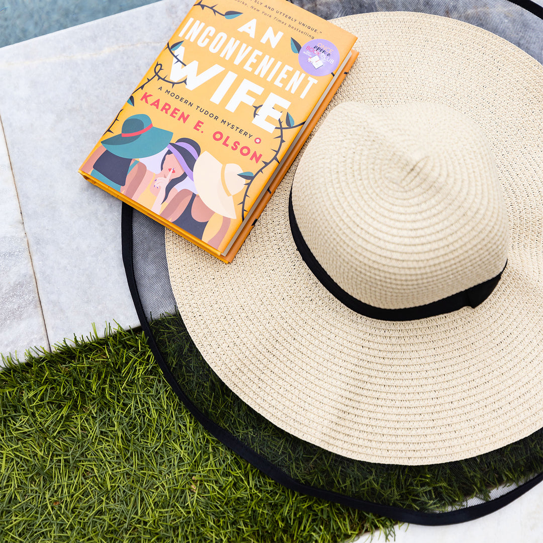 a cream colored sunhat with a mesh edge and black ribbon. a copy of An Inconvenient Wife by Karen E. Olson sits next to the hat.