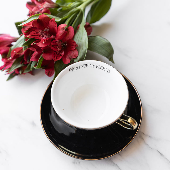 A black tea cup and saucer sits on a white marble counter. The teacup is white on the inside and features a gold handle. The quote "You Stir My Blood" is printed on the inside of the cup.