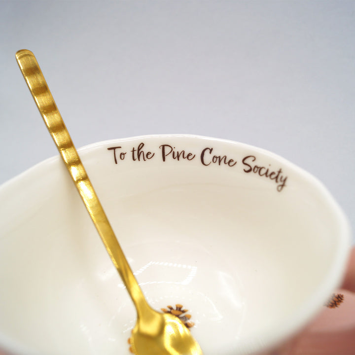 A closeup of the inside of the teacup, printed with the words "To the Pine Cone Society" and the gold teaspoon inside the cup.