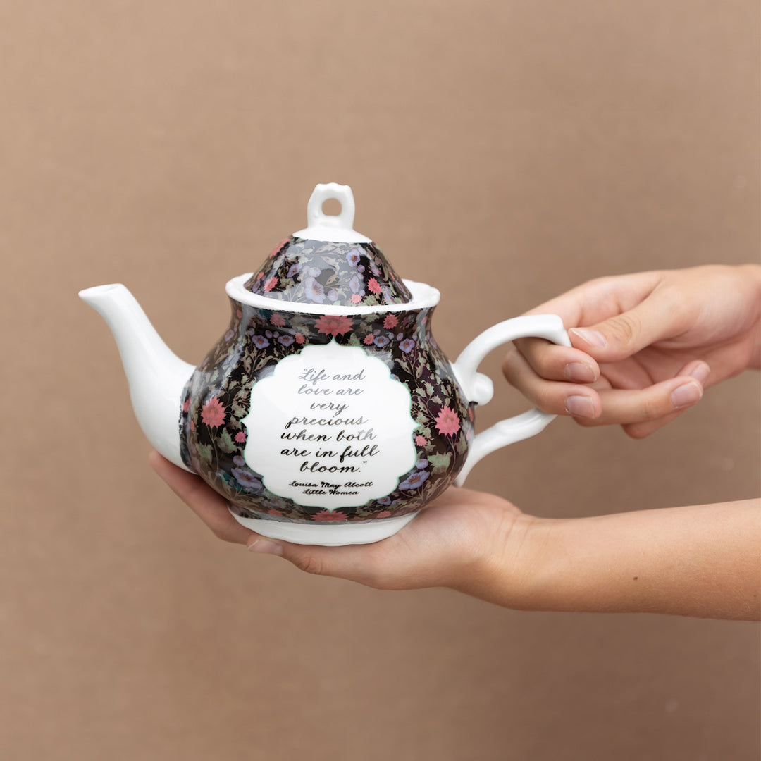 Hands hold out a ceramic teapot with a black floral pattern on the body and top while the spout and handle are white. A white circular area is in the middle of the black floral area with the quote: Life and love are very precious when both are in full bloom. Louisa May Alcott Little Women.