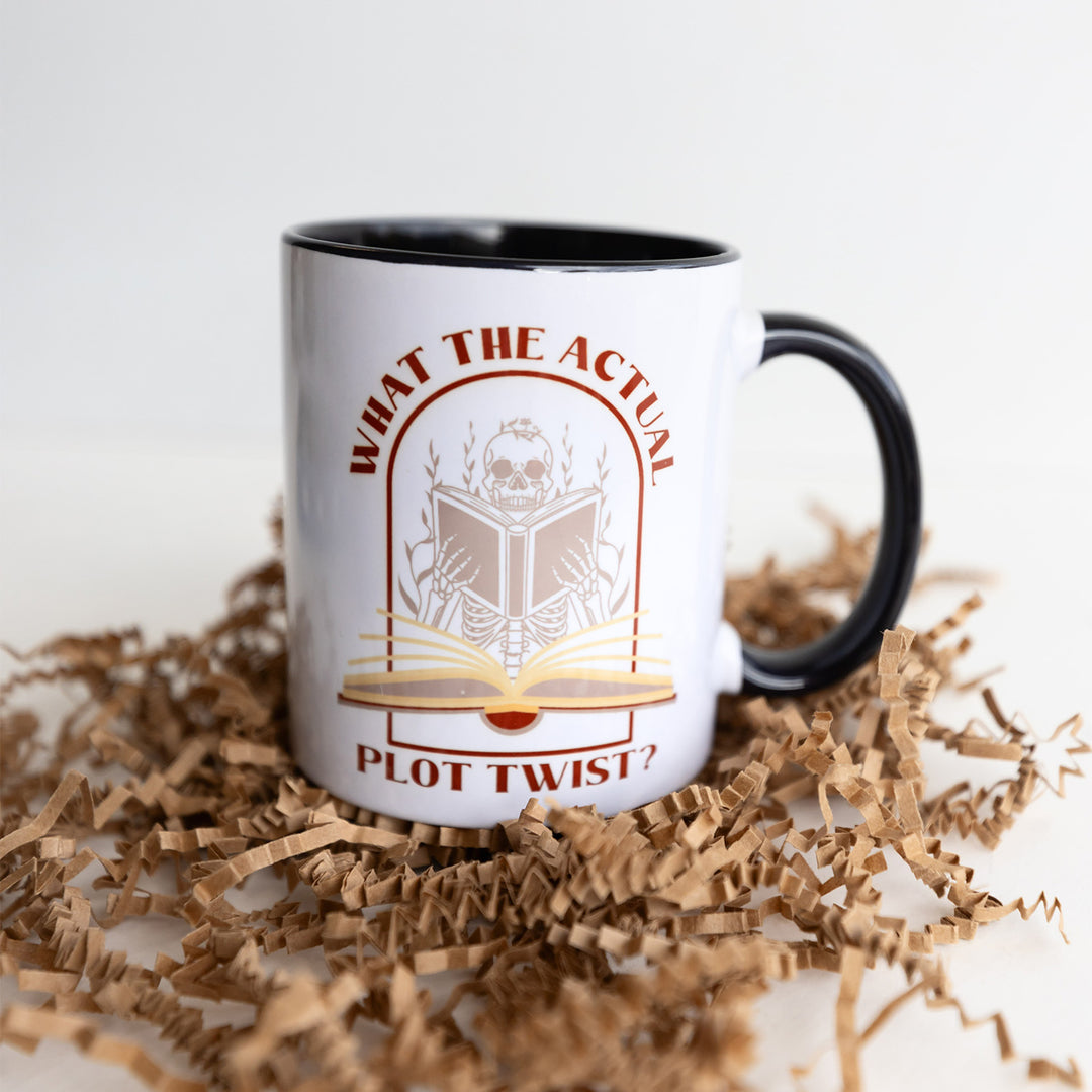 A white mug with a black handle and black inside, with the quote What the Actual Plot Twist? on the side with an image of a skeleton reading a book.
