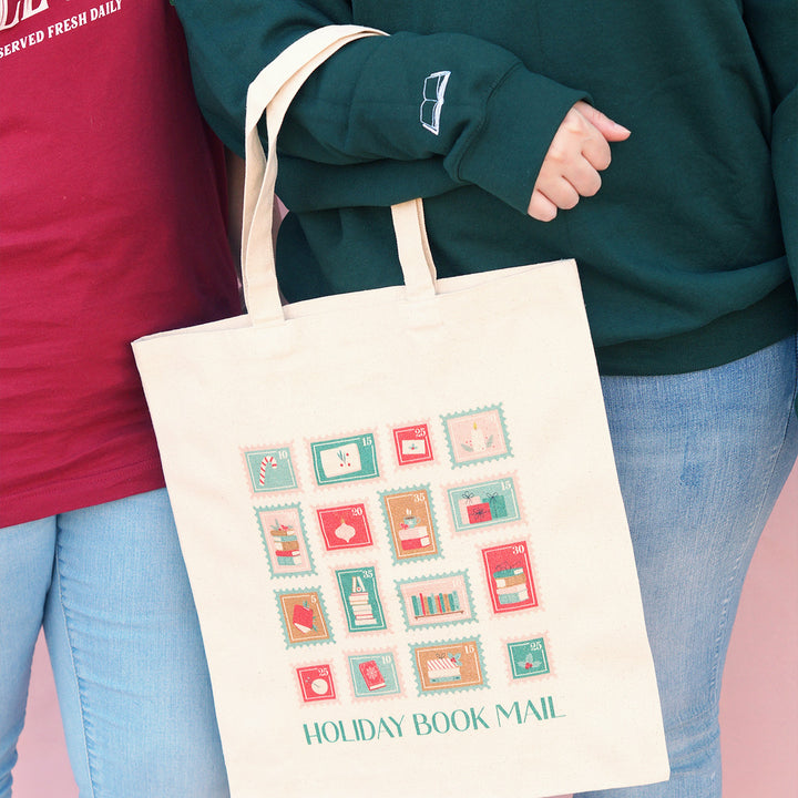 Tote bag with 'Holiday Book Mail' stamp design held by a person wearing a green sweater.