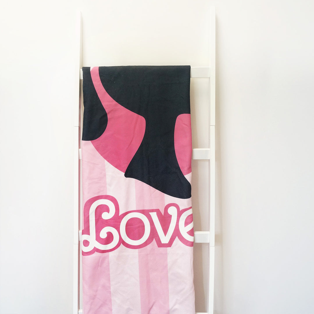 A pink towel hanging over a blanket ladder. The towel has light pink stripes on it with the word Love and a black silhouette of a woman's face profile on it.