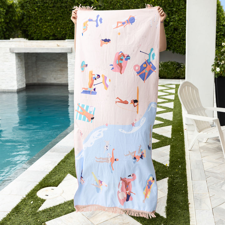 A beach scene themed beach towel being held up by a pair of hands next to an in-ground pool. The top of the towel features a light pink beach with multiple people enjoying the sun and good books. The bottom of the towel features a light blue ocean with surfers and swimmers enjoying the waves.