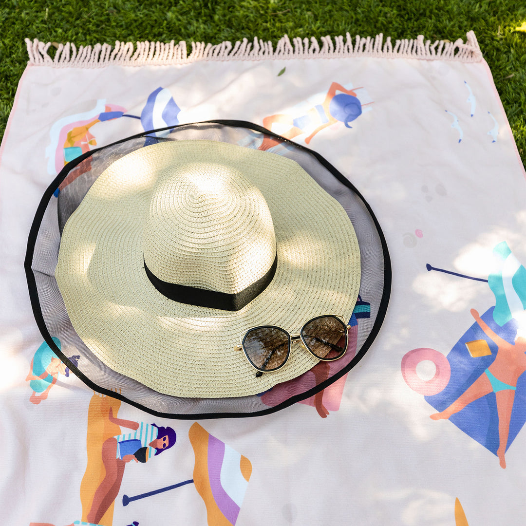 A close up of part of the beach scene beach towel. A cream and black sun hat and pair of sunglasses sits on the towel. People in the illustrated beach scene on the towel sit on the beach with their books and enjoy the sun. Pink tassels stick out from the top of the towels edge in contrast to the green grass.