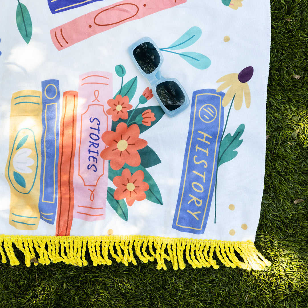 A close-up of the bottom right corner of the book-themed beach towel. Book spines reading "Stories" and "History" are visible. Red and yellow flowers and green leaves are stacked between the books. A yellow book has a white flower printed on the spine. A pair of blue sunglasses face the camera. The yellow tassels at the bottom of the towel are bold against the green grass.