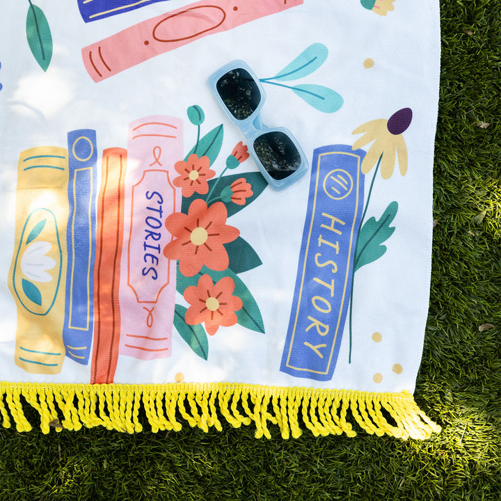 A close-up of the bottom right corner of the book-themed beach towel. Book spines reading "Stories" and "History" are visible. Red and yellow flowers and green leaves are stacked between the books. A yellow book has a white flower printed on the spine. A pair of blue sunglasses face the camera. The yellow tassels at the bottom of the towel are bold against the green grass.