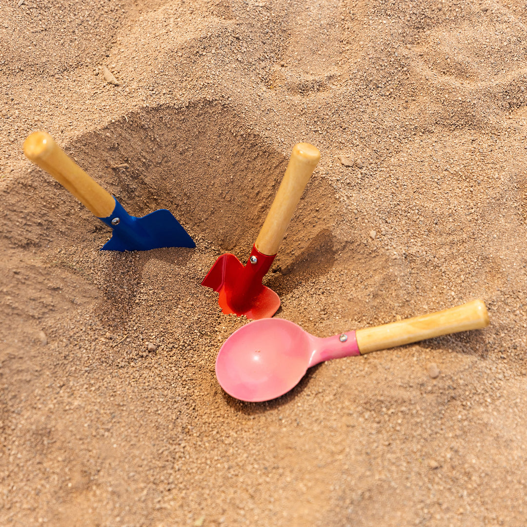 Three shovel/scoop tools lay in the sand - one blue, one red, and one pink, all with wooden handles
