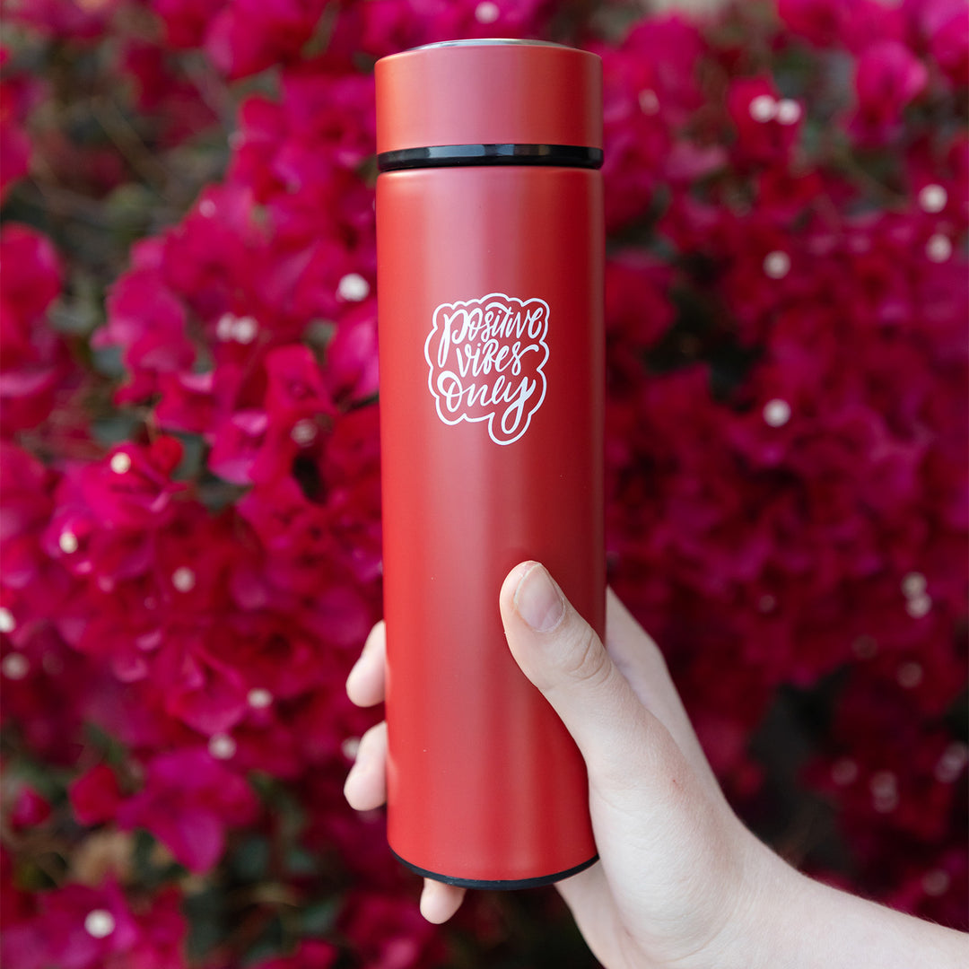 A white hand holds a red tumbler in front of pink flowers. The red tumbler says Positive Vibes Only in white lettering.