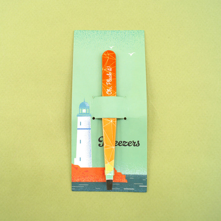 Orange and yellow ombre tweezers that say Oh, Pluck it! on the side. They are attached to a blue piece of cardboard with a lighthouse and the ocean on it.