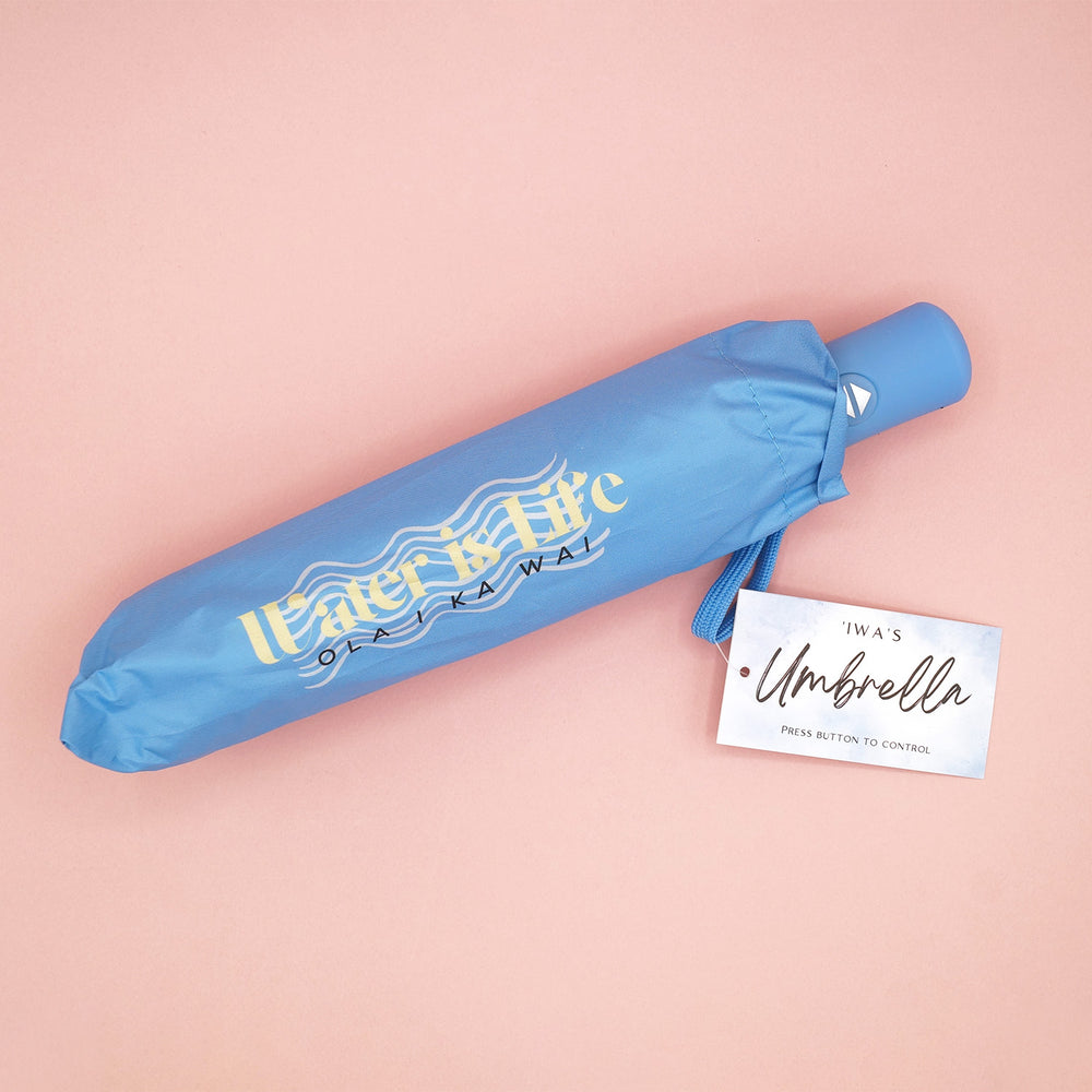 A blue umbrella that says Water is Life Ola Ika Wai on it with a wave pattern behind the words.