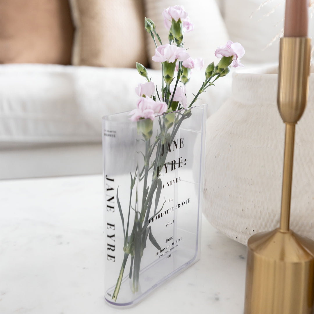 A clear acrylic book-shaped vase printed to look like a copy of Jane Eyre by Charlotte Bronte sits on a white surface next to other home decor. The vase holds a small bouquet of pink flowers.