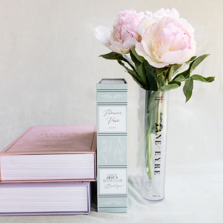 A clear acrylic book-shaped vase printed to look like a copy of Jane Eyre by Charlotte Bronte sits on a white surface, holding a bouquet of pink and white flowers. The spine of the book vase faces the camera directly. Next to the vase is the green box the vase is packed inside upon purchase.