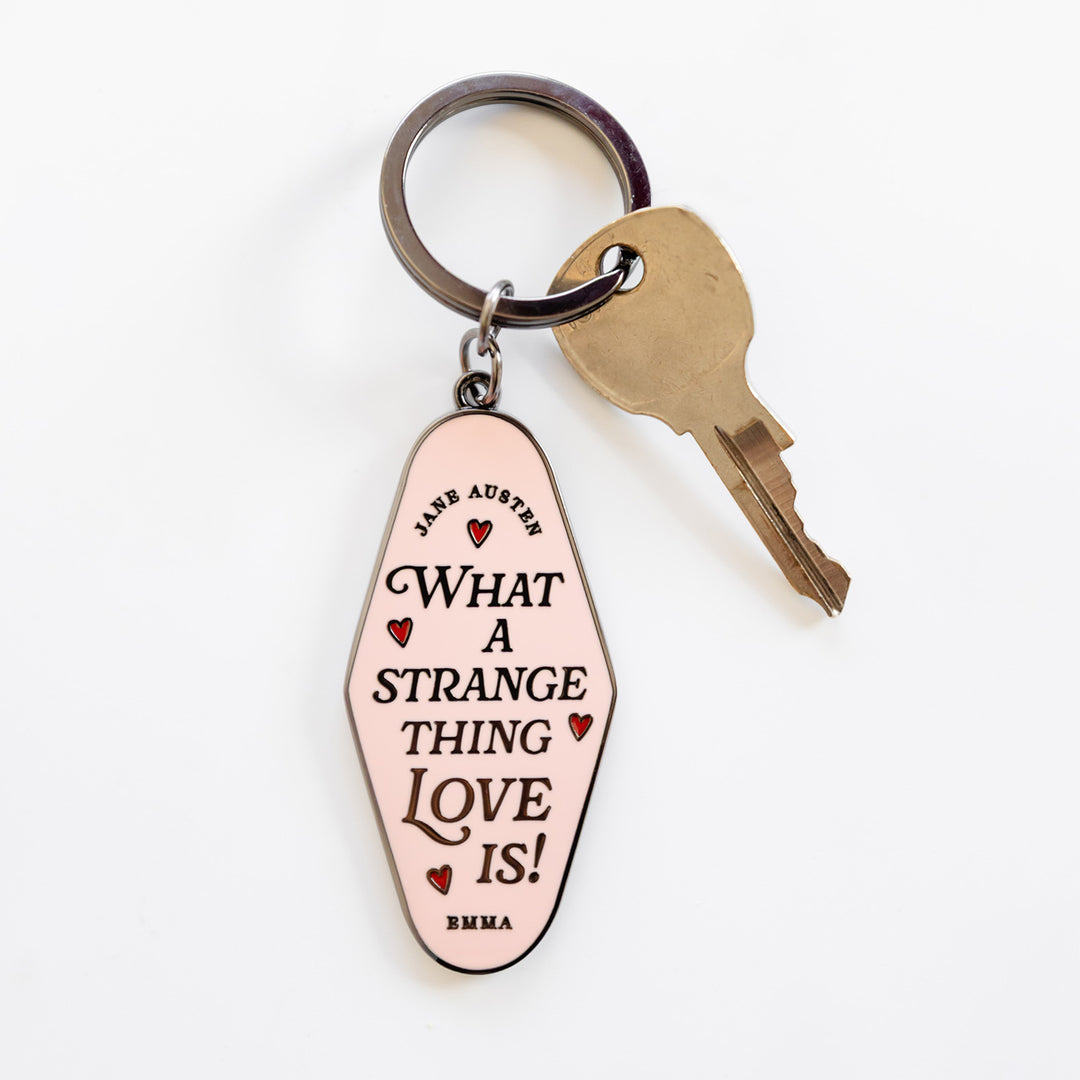 A key ring with a gold key and a light pink keychain. The keychain has four small red hearts surrounding the black text in the middle: Jane Austen 'What a Strange Thing Love Is!' Emma