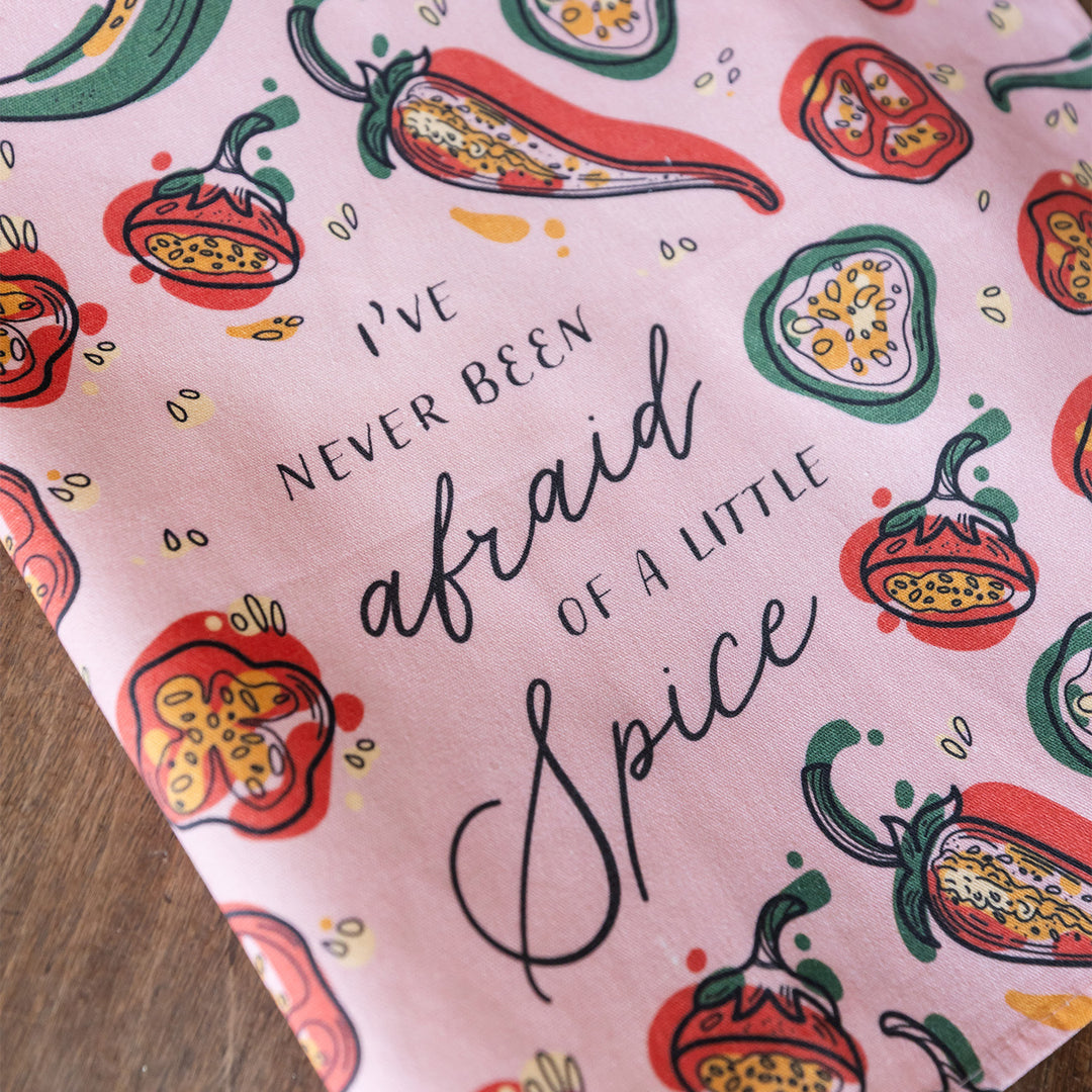 A light pink kitchen towel with red and green chili pepper pattern. In black text is the quote: I've Never Been Afraid of a Little Spice.