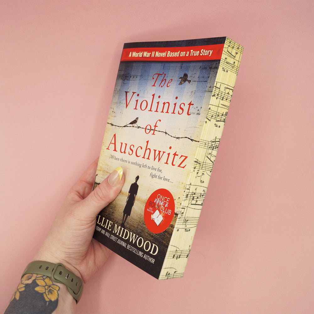 Book titled 'The Violinist of Auschwitz' by Ellie Midwood showcasing patterned page edges is held by a hand on a pink background