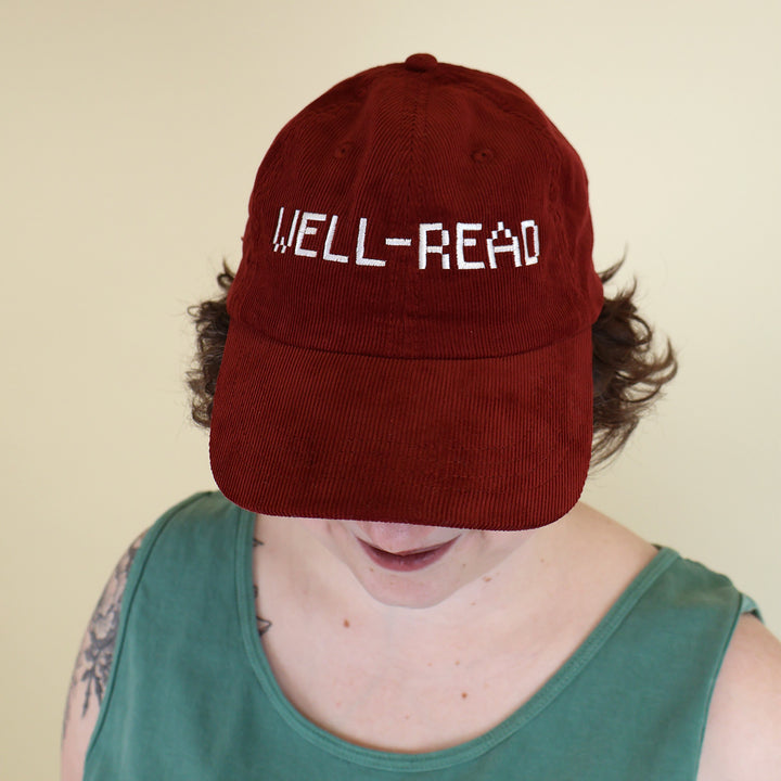 Woman wearing a maroon corduroy cap embroidered with 'WELL-READ' text on a beige background