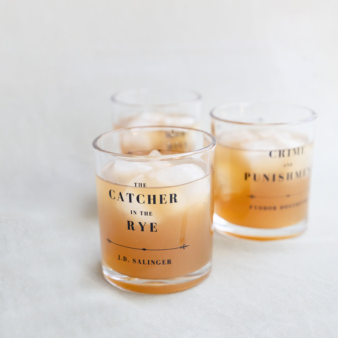 A set of whiskey glasses inspired by classic book titles. A close up on The Catcher in the Rye by J.D. Salinger glass with Crime and Punishment by Fyodor Dostoevsky visible behind it. Each glass is filled with an amber liquid.