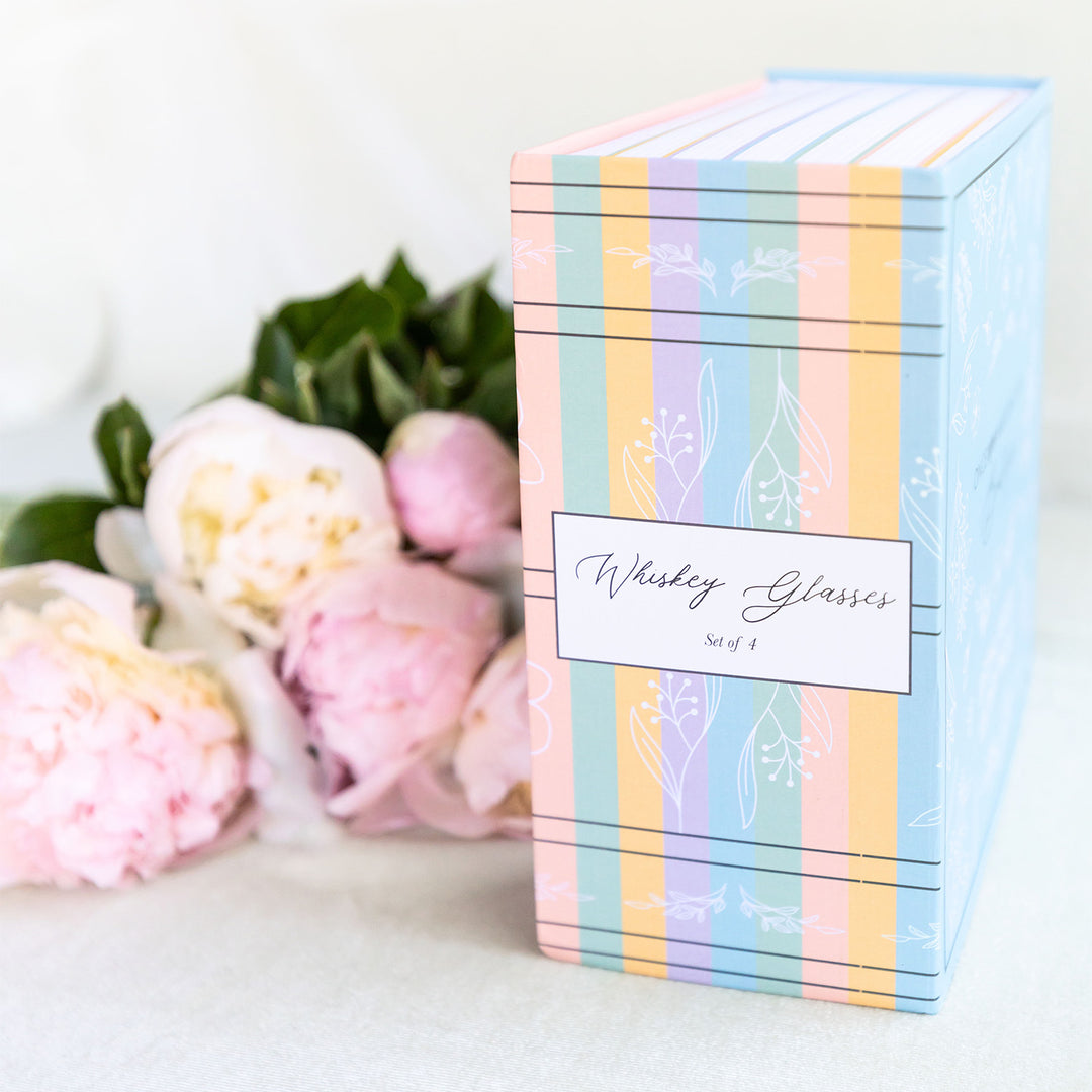 The packaging box for the whiskey glasses. This box is multicolored and appears to have many book spines along its edge. The box reads Whiskey Glasses Set of 4. Pink flowers lay next to the box on the left.