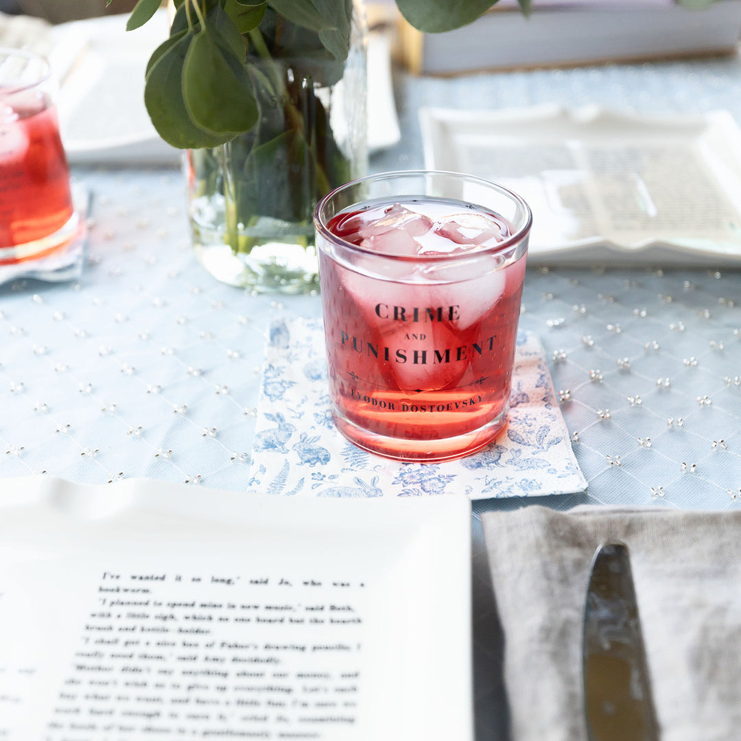 A set of whiskey glasses inspired by classic book titles. This close up of Crime and Punishment by Fyodor Dostoevsky shows it filled with a red beverage and ice. A table setting sits in front of the glass. A bouquet of flowers sits behind it on the table.