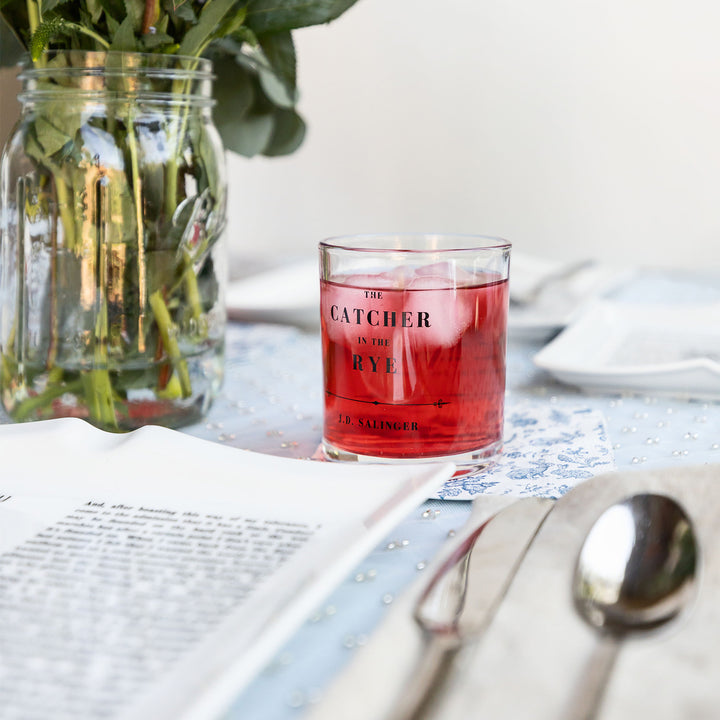 A set of whiskey glasses inspired by classic book titles. This close up on The Catcher in the Rye by J.D. Salinger glass shows it filled with a red beverage and ice. A table setting sits in front of the glass. A bouquet of flowers sits next to the glass.