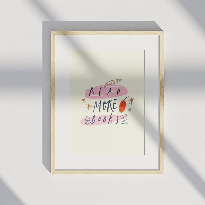 A matted wooden frame hangs on a white wall and displays the "Read More Books" art print. A shadow from a window is visible over the art print.