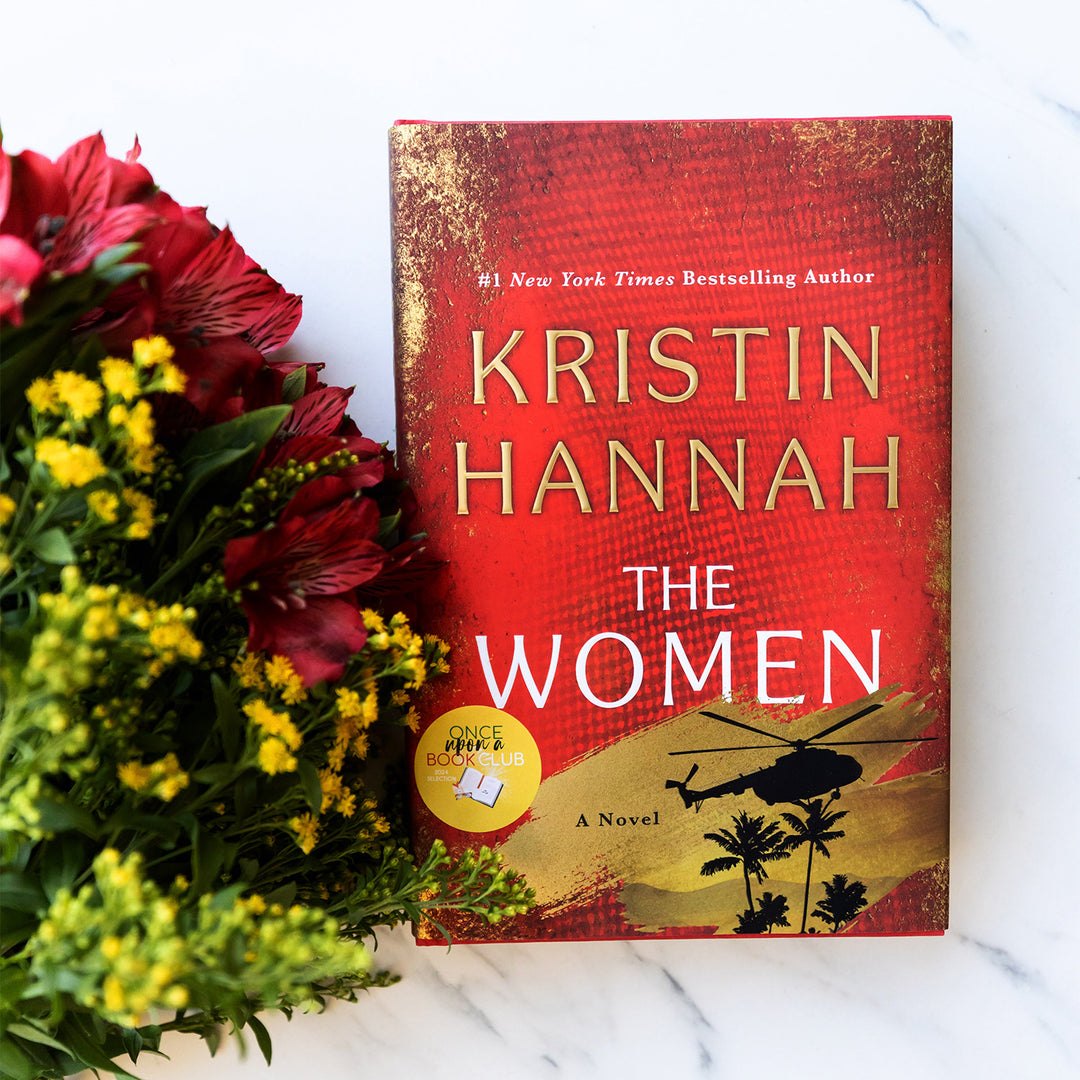 A hardcover copy of The Women by Kristin Hannah sits on a white background next to a bouquet of red flowers with yellow smaller flowers.