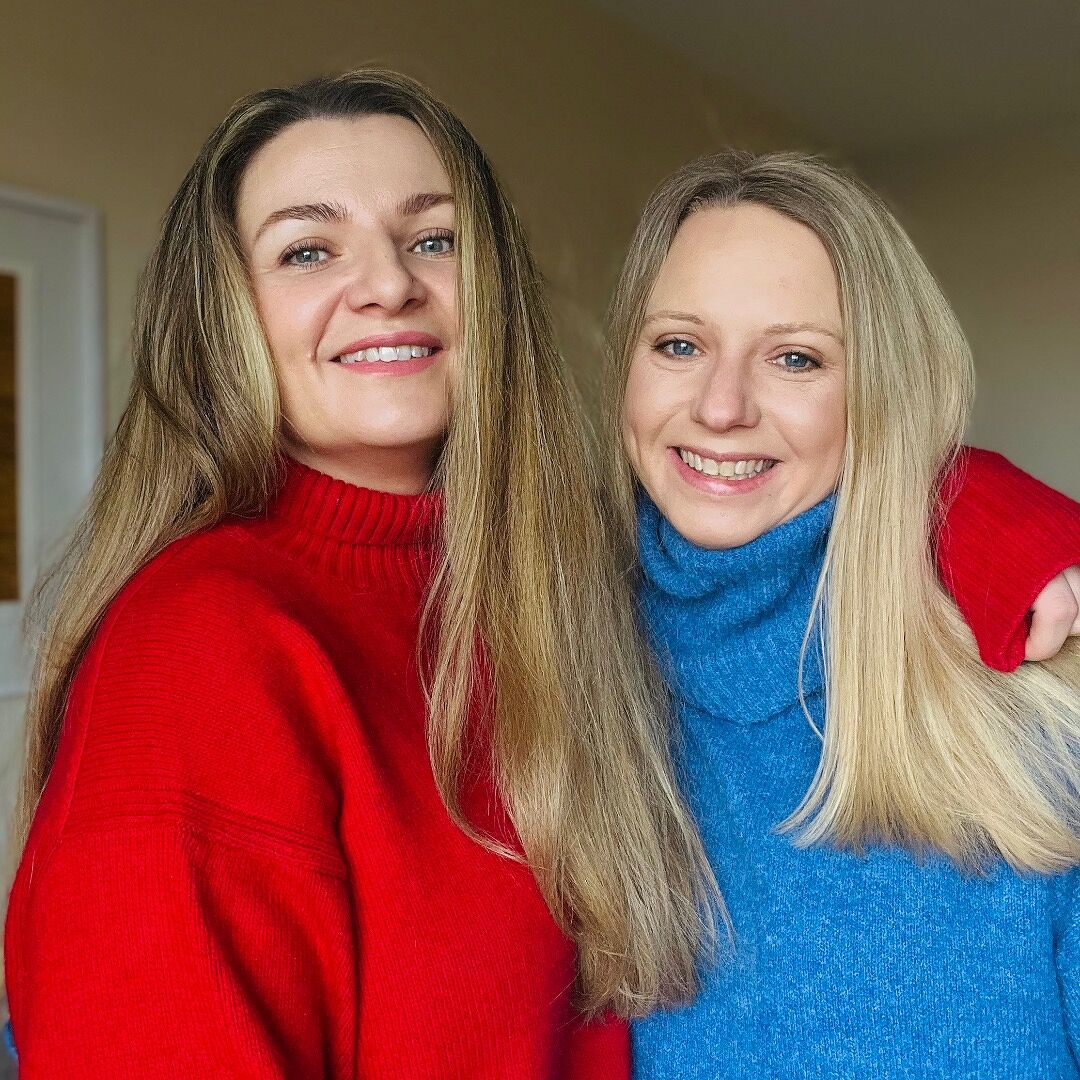 Ellis Hunter, two women smiling and posing together indoors, wearing bright red and blue turtleneck sweaters. The woman on the left wears a red sweater, and the woman on the right wears a blue sweater, with their arms around each other.