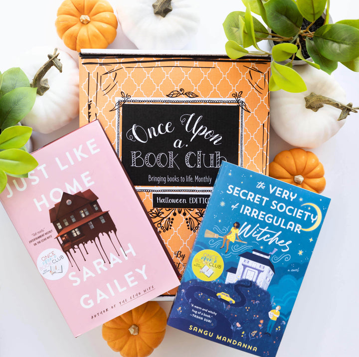 a hardcover edition of Just Like Home by Sarah Gailey and a paperback edition of The Very Secret Society of Irregular Witches by Sangu Mandanna lay on an orange Once Upon a Book Club Halloween box surrounded by orange and white pumpkins