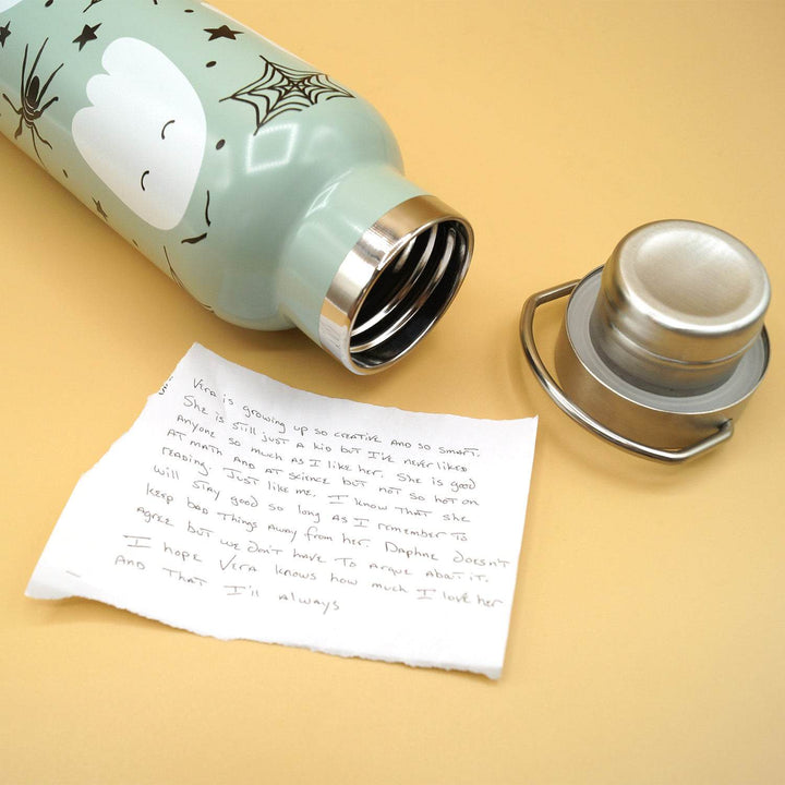 a light green water bottle lays open on its side with a pattern of white ghosts and black spiderwebs next to the silver lid and white scrap of paper with handwriting on it