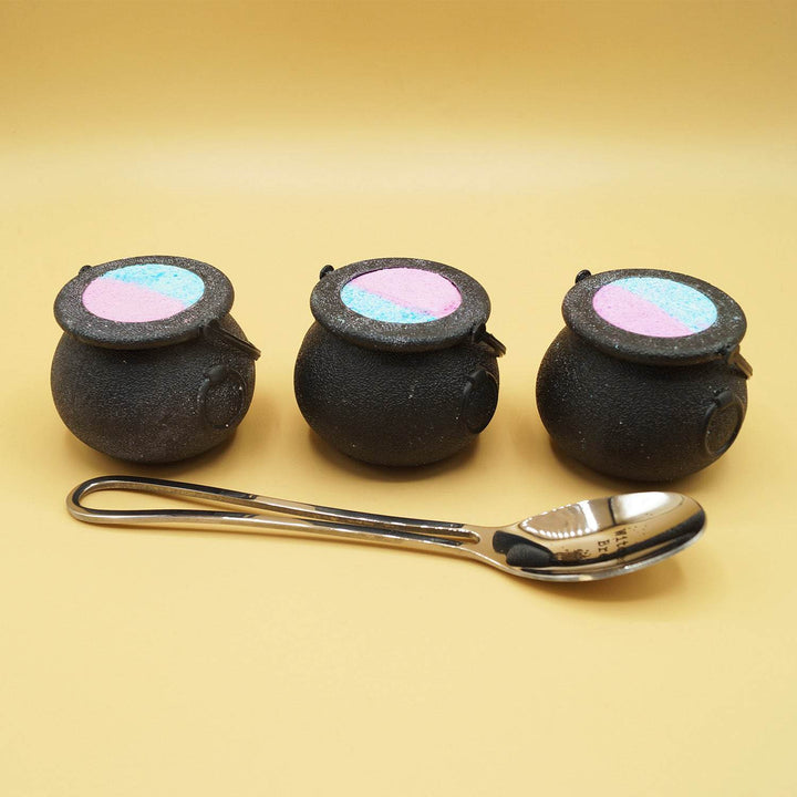 3 black cauldron-shaped bath bombs stand in a line behind a silver spoon